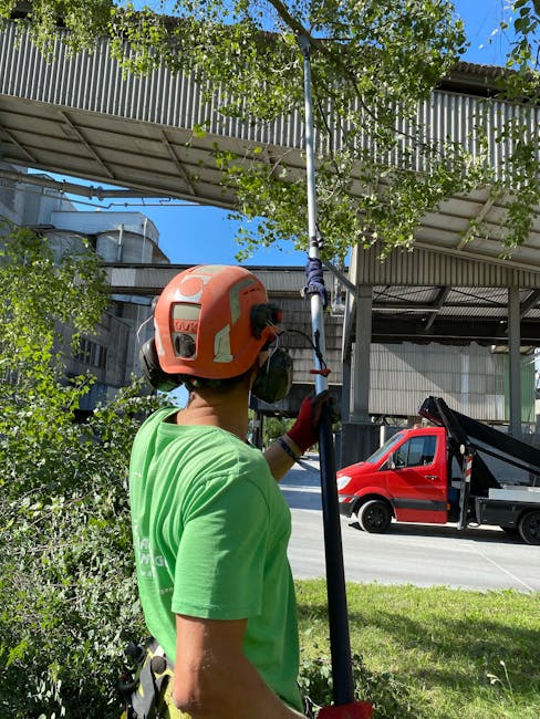A gardener wearing a bright green t-shirt and an orange safety helmet is engaged in tree maintenance, holding a pole with a pruning or cutting tool at the end. The background features a modern industr